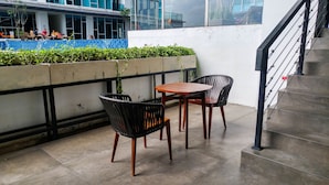 A balcony with two wicker chairs and a small table, a concrete floor, and a view of a building with people sitting outside. - Minimalist Style Studio Brooklyn Apartment near IKEA Alam Sutera (South Tangerang)