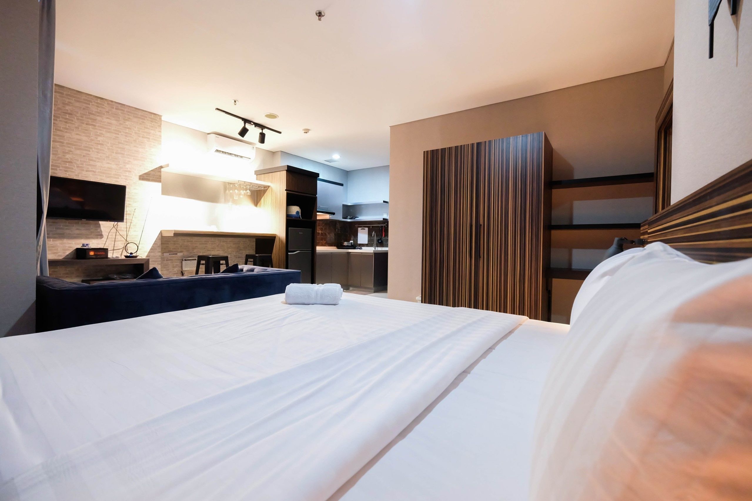 Une chambre d’hôtel moderne dotée d’un grand lit, d’un téléviseur à écran plat fixé au mur, d’un coin cuisine intégré et d’un mur revêtu de panneaux en bois.