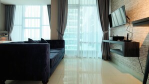 A modern living room with a dark sofa, a flat-screen TV mounted on a brick wall, and a large window with sheer curtains. - Spacious and Artsy Studio Apartment at Brooklyn Alam Sutera (South Tangerang)
