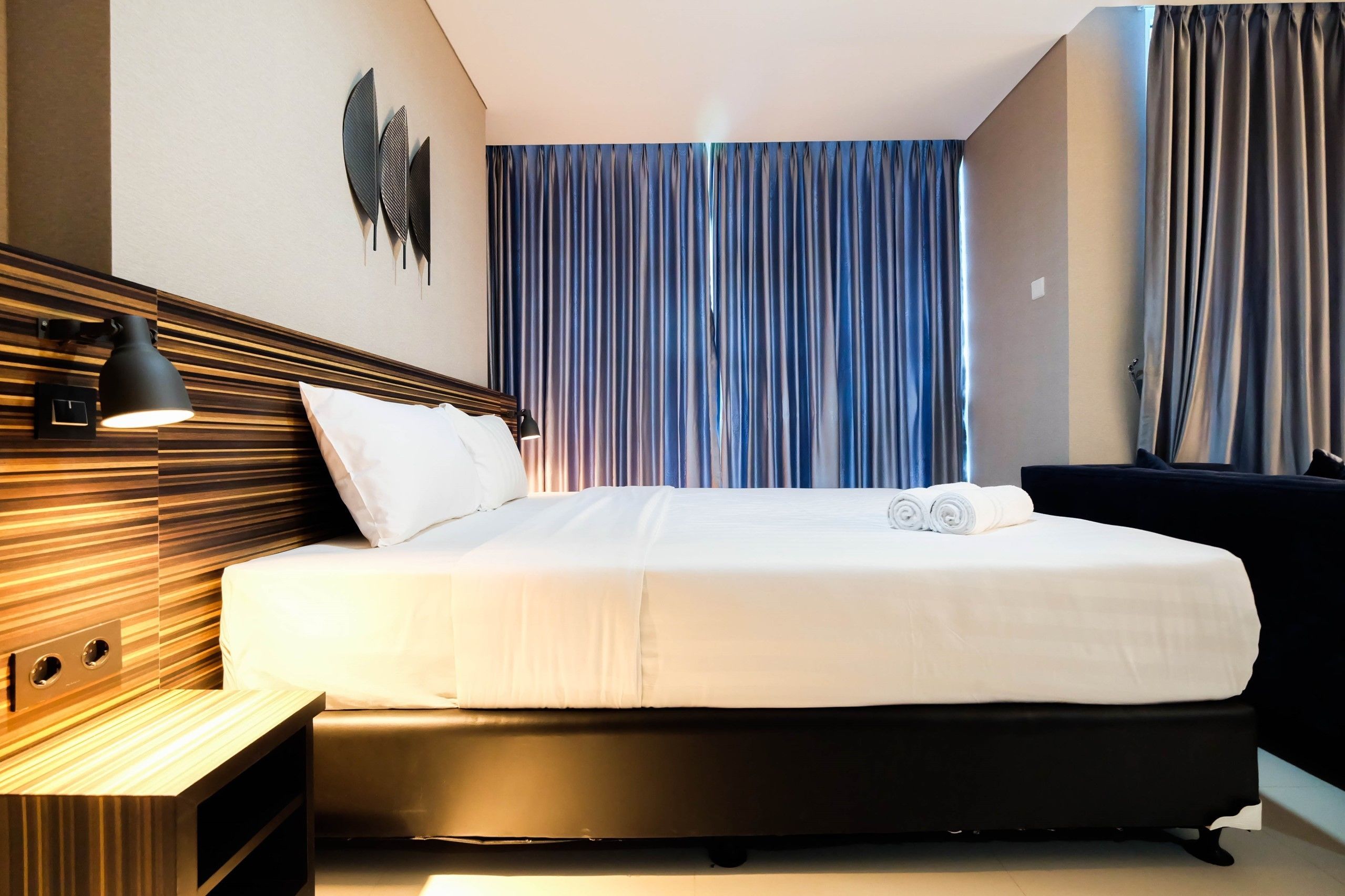 A modern hotel room with a large bed, a bedside lamp, a wall-mounted decorative piece, and a view of the outside through curtains.