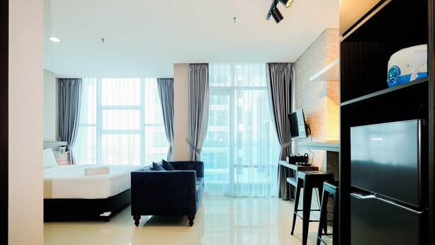 A modern hotel room with a bed, a sofa, a TV, a refrigerator, and a kitchen area. - Spacious and Artsy Studio Apartment at Brooklyn Alam Sutera (South Tangerang)