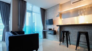 A modern living room with a sofa, a flat-screen TV mounted on a brick wall, and a bar area with stools. - Spacious and Artsy Studio Apartment at Brooklyn Alam Sutera (South Tangerang)