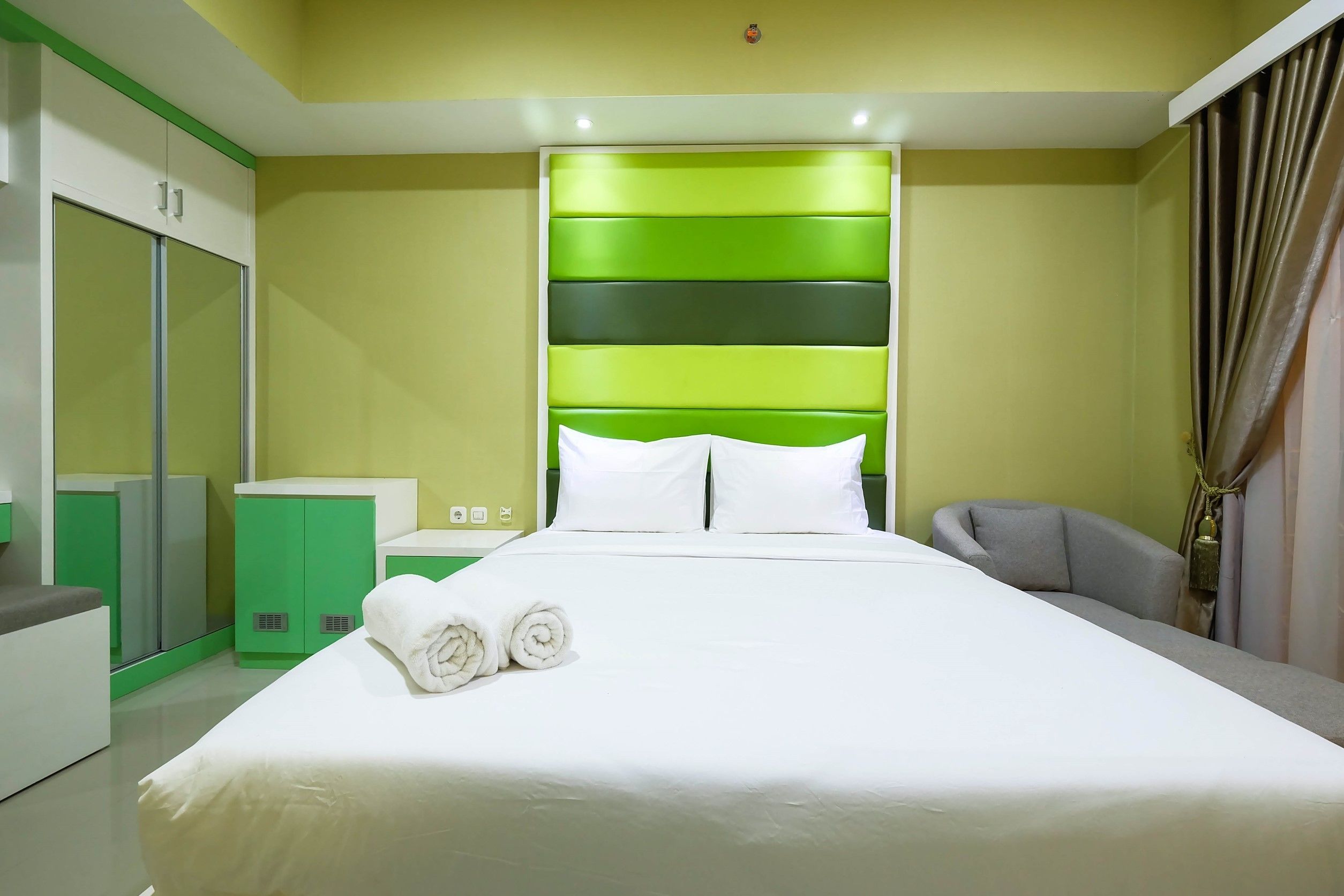 Une chambre d’hôtel moderne avec un grand lit, une armoire verte et blanche, et un fauteuil gris.