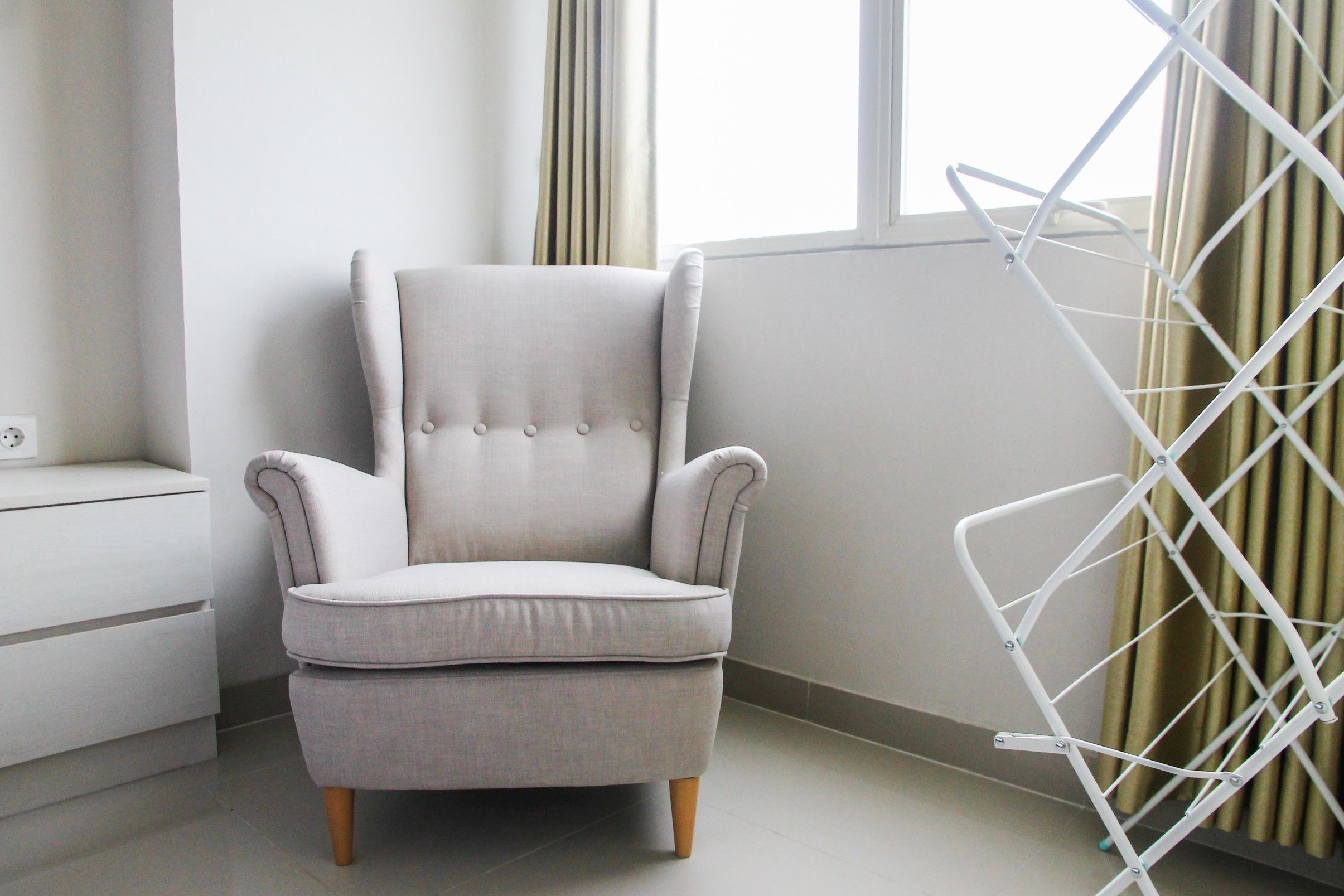 A light grey upholstered armchair with wooden legs, positioned next to a white radiator and a white drying rack.
