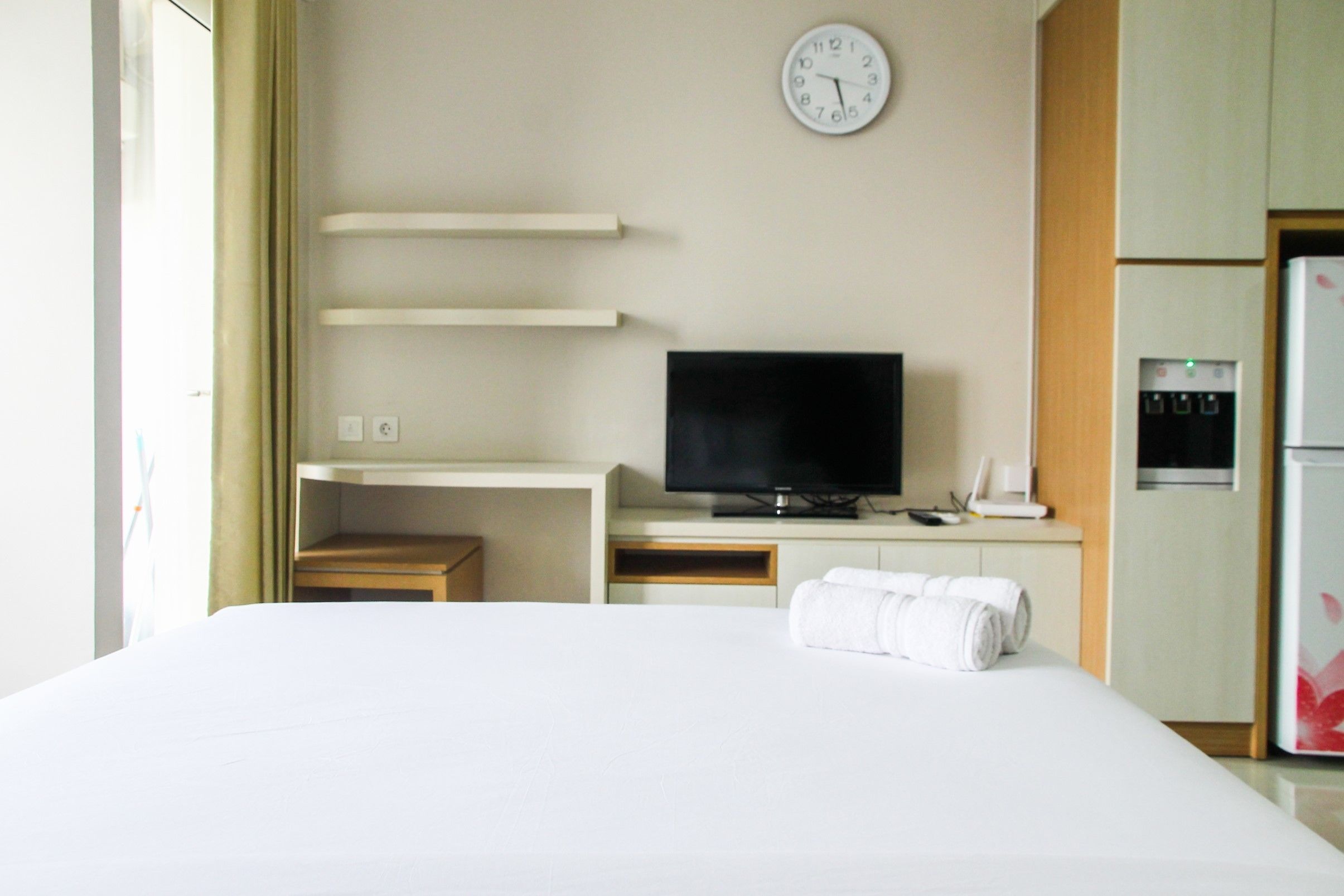 A hotel room with a bed, a flat-screen TV, a clock, a water dispenser, and a small table.