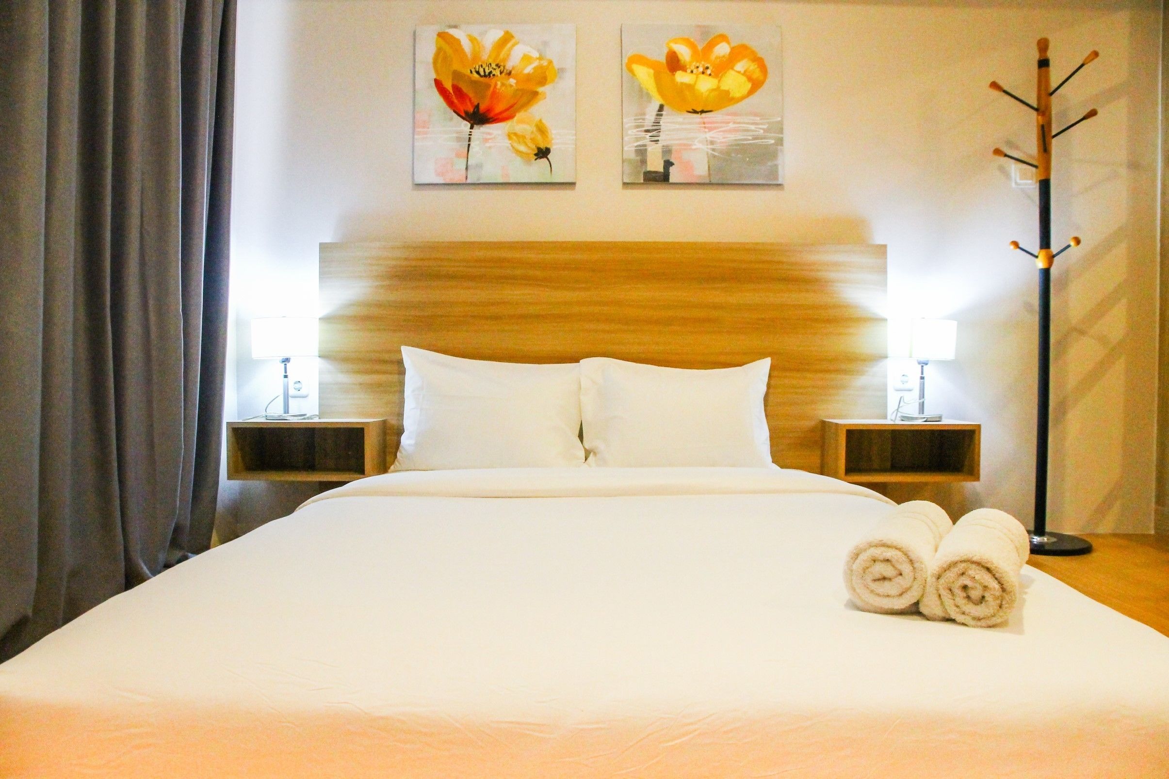 Una cama bien tendida con sábanas blancas, dos cuadros de flores colgados en la pared, un cabecero de madera y una mesita de noche con una lámpara.