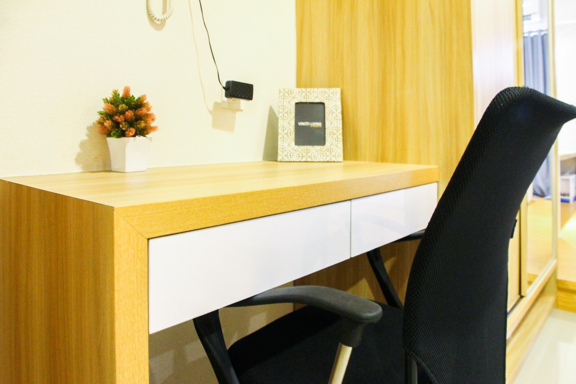 A wooden desk with a white drawer, a black office chair, a small potted plant, and a picture frame on the wall.
