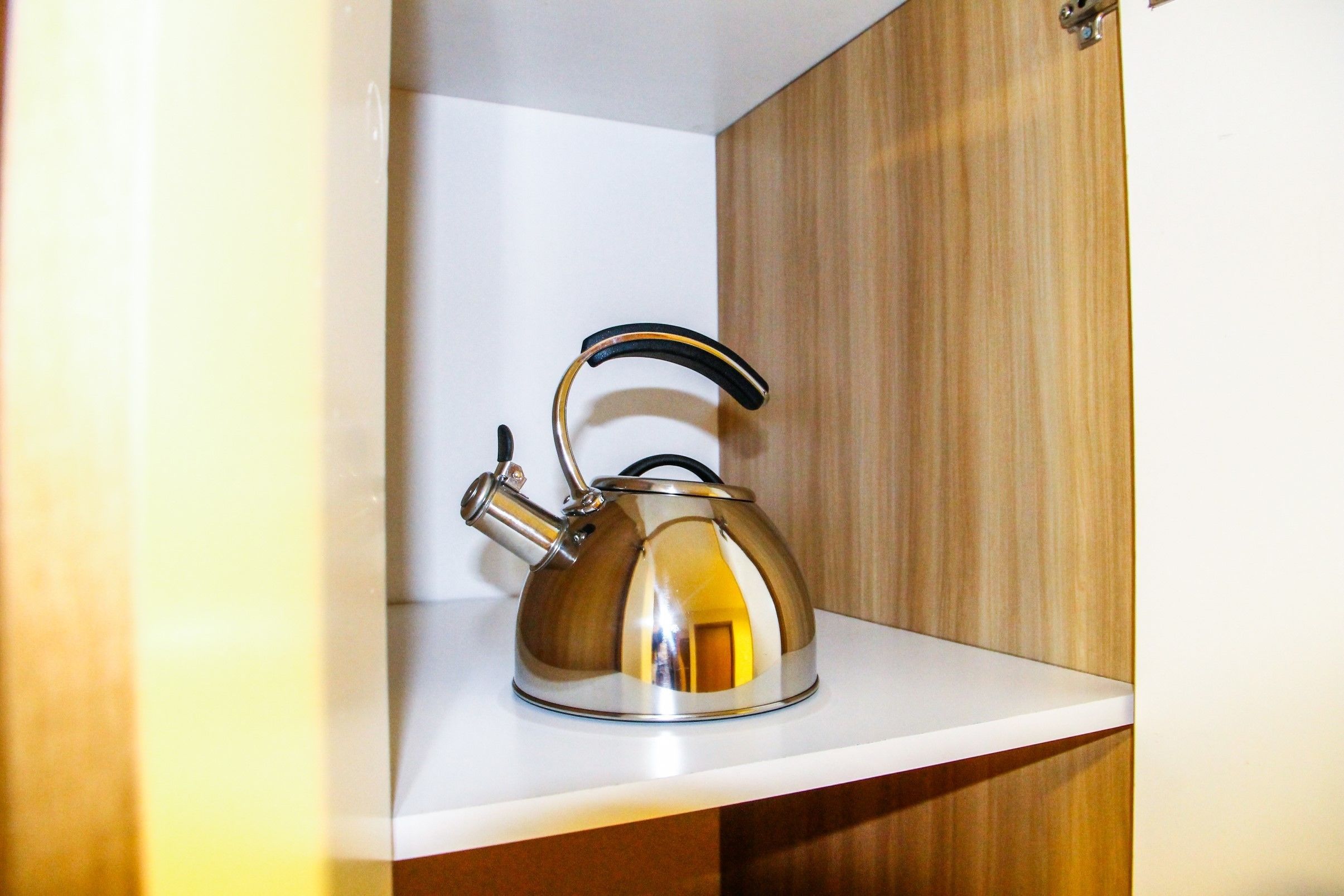A stainless steel kettle on a white shelf in a wooden cabinet.