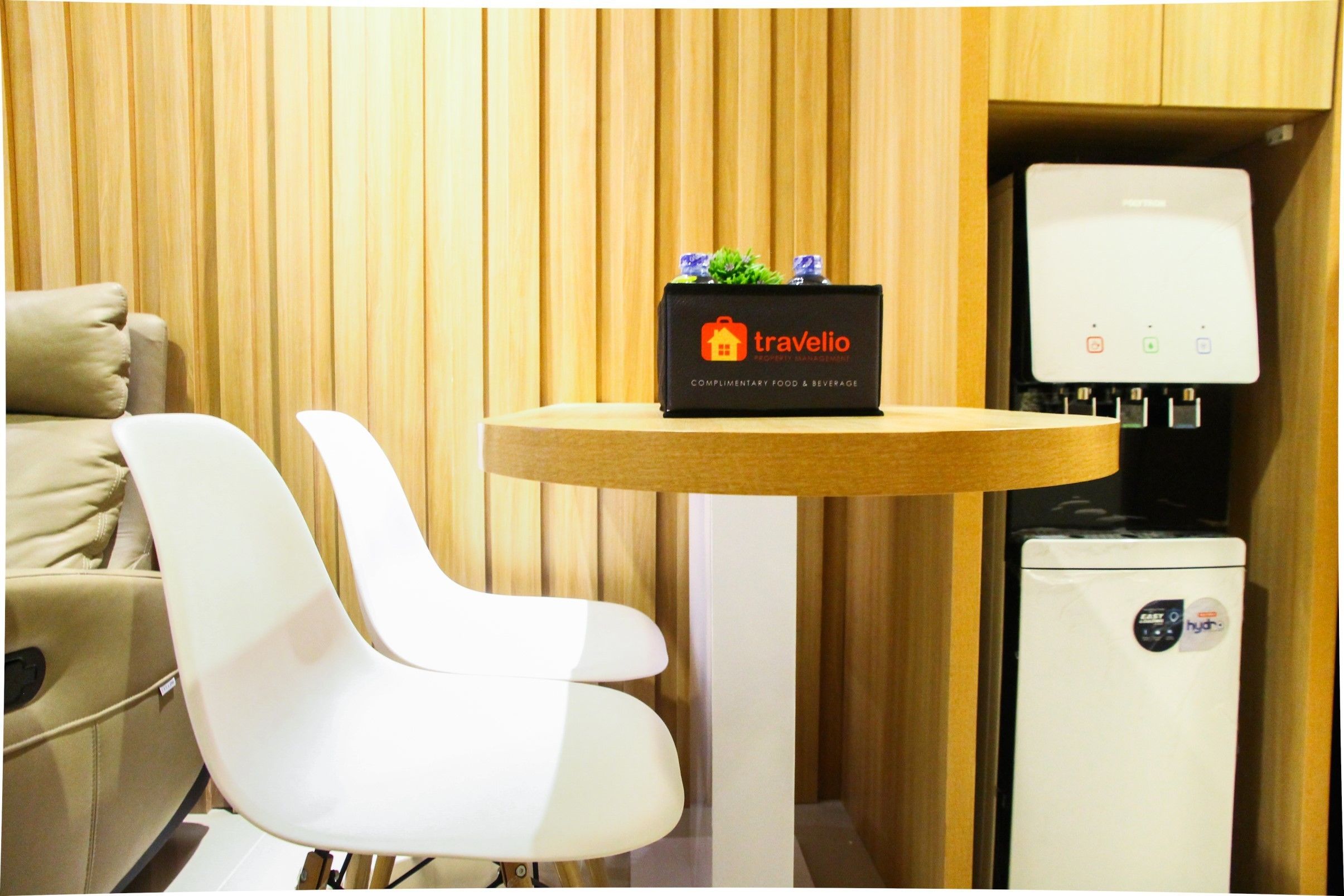 A modern hotel room with a wooden panel wall, a white chair, a small round table with a 'Travelio' sign, and a mini-fridge.