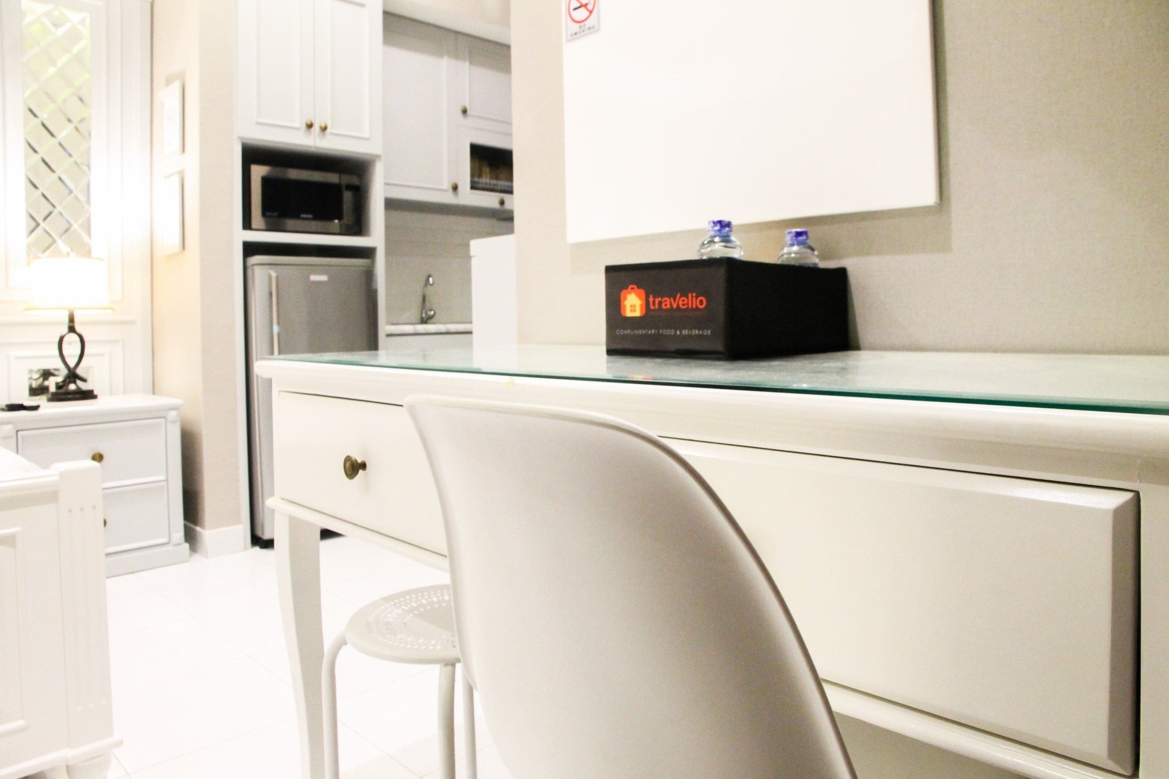 Une chambre d’hôtel moderne avec un bureau à plateau en verre, une chaise blanche et un mini-bar.