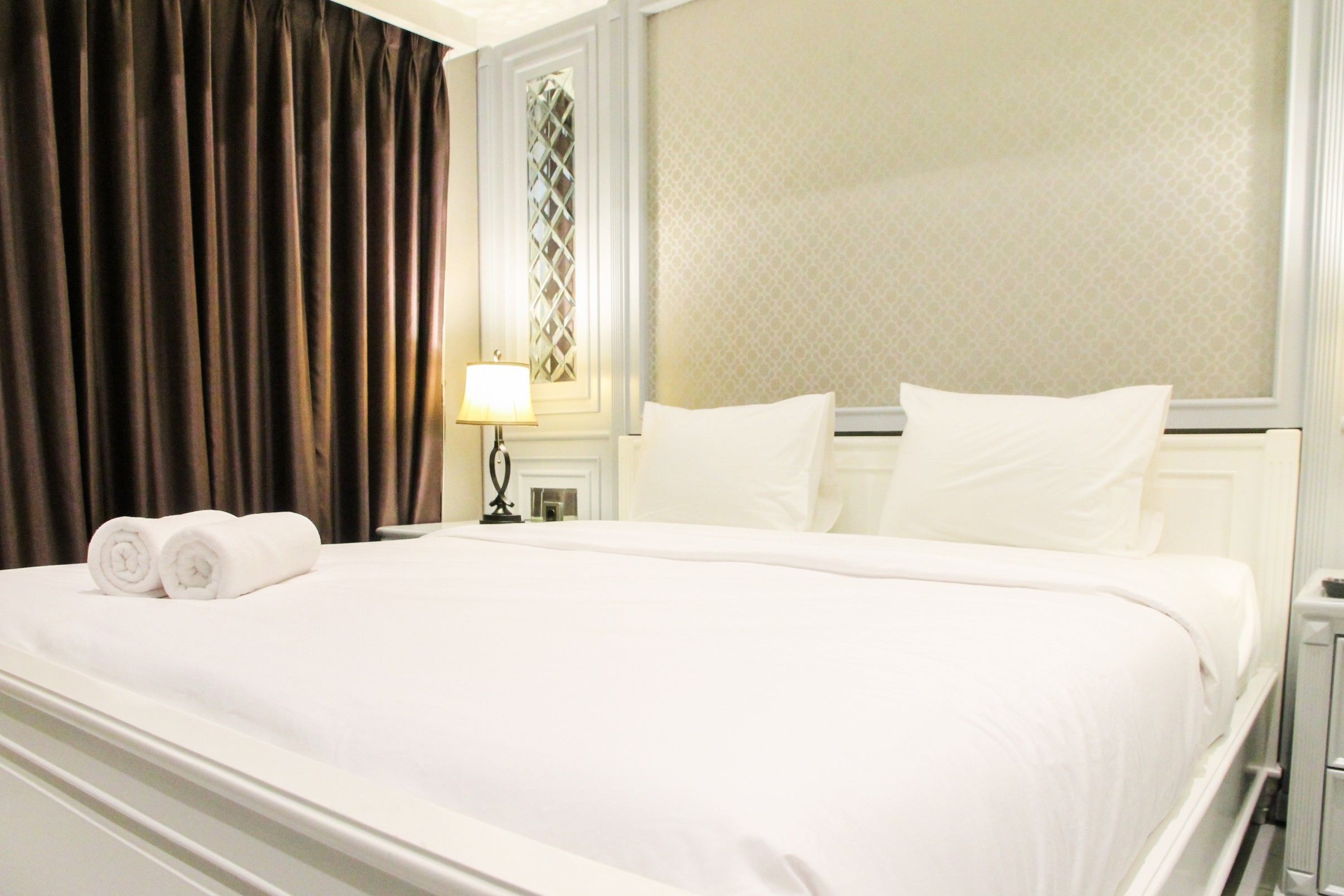 A hotel room with a large bed, two pillows, a nightstand with a lamp, and a window with curtains.