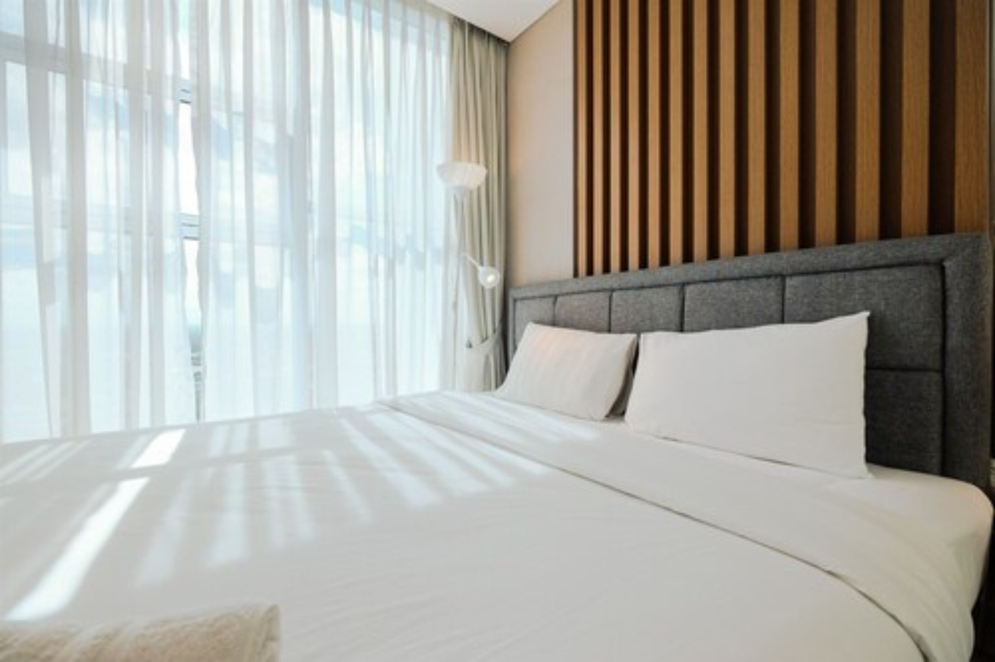 Quarto de hotel moderno com uma cama grande, parede de painéis de madeira e uma janela ampla com cortinas leves.
