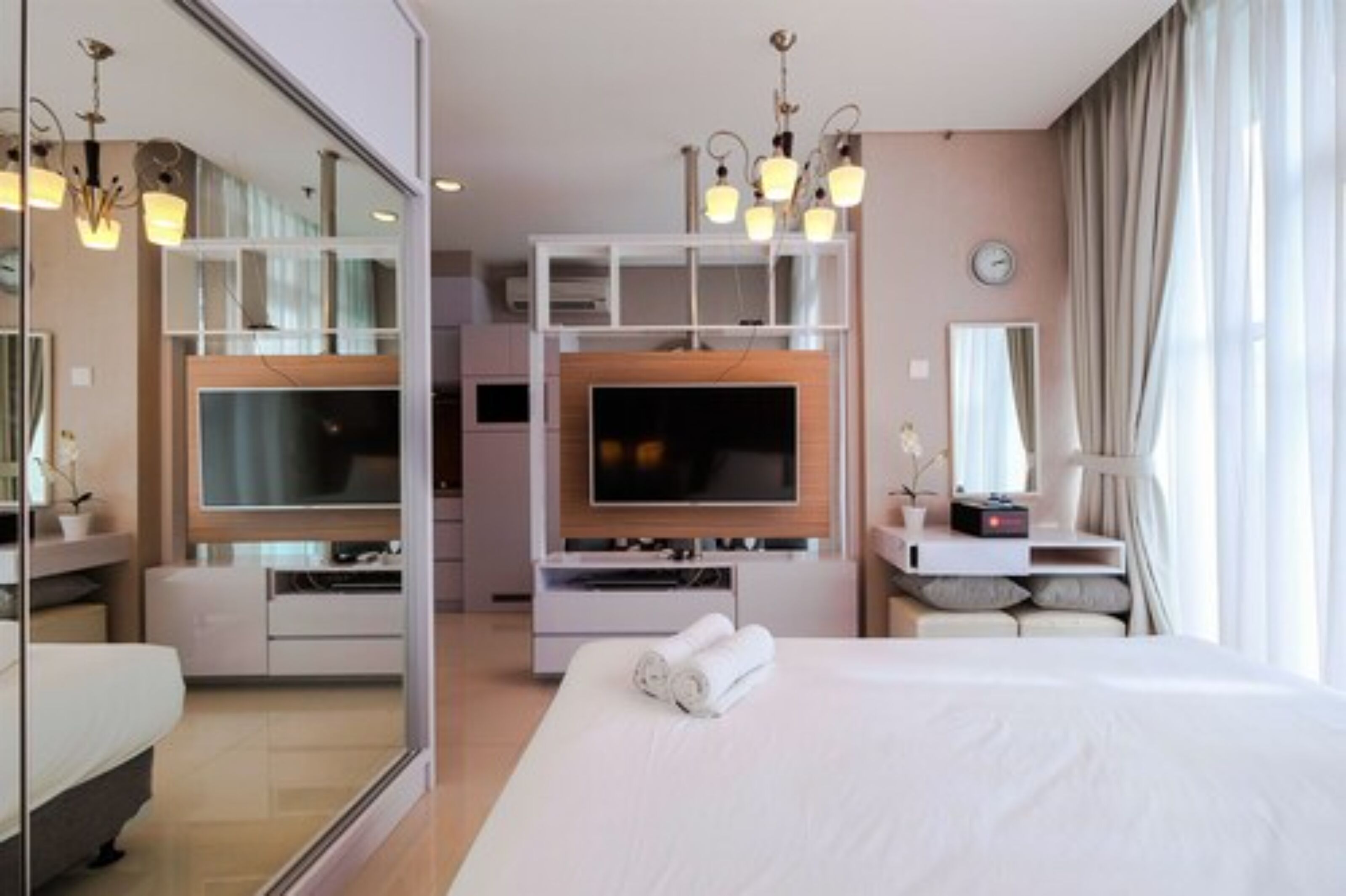 A modern hotel room with a large bed, a flat-screen TV mounted on a wooden panel, a bedside table with a lamp, and a mirror above the bed.