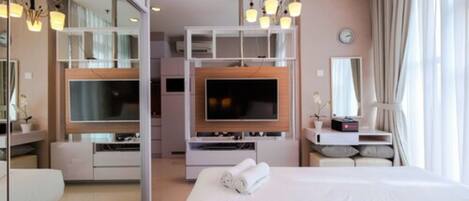 A modern hotel room with a large bed, a flat-screen TV mounted on a wooden panel, a bedside table with a lamp, and a mirror above the bed.