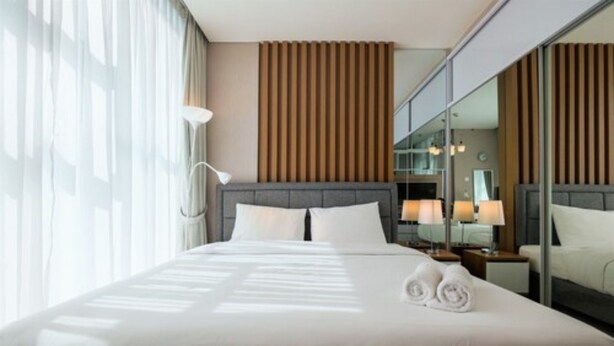 A modern hotel room with a large bed, a nightstand with lamps, a mirror, and a view of the city through floor-to-ceiling windows.