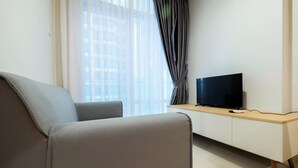 A modern living room with a grey sofa, a flat-screen TV on a wooden console, and sheer curtains on a large window. - Well Equipped 1BR Brooklyn Alam Sutera Apartment near IKEA (South Tangerang)