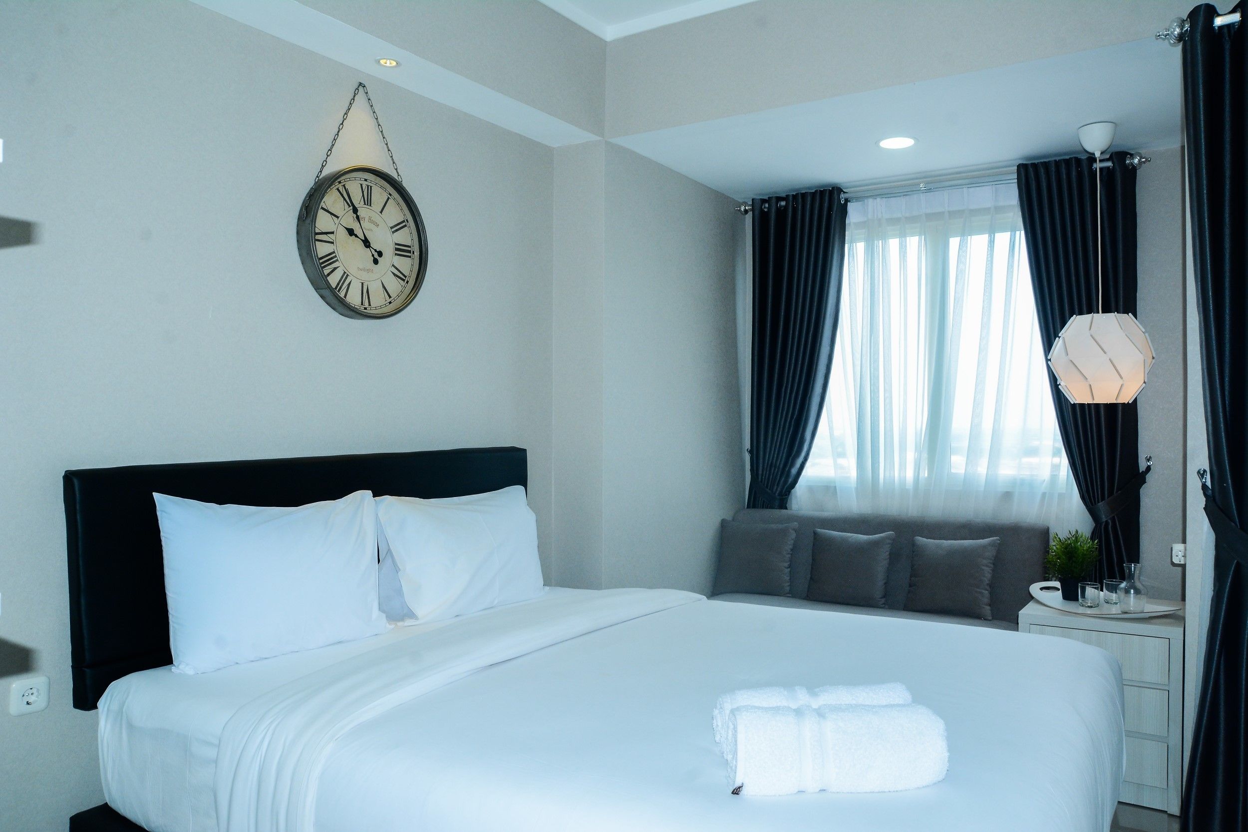 A neatly made bed with white linens and a folded towel, a round wall clock, and a window with dark curtains.