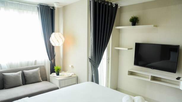 A hotel room with a bed, a TV, a sofa, a nightstand, and a window with curtains. - Sophisticated Studio at The Oasis Apartment (Cikarang)