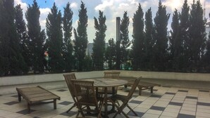 An outdoor seating area with a tiled floor, surrounded by tall trees and a tall building in the background.