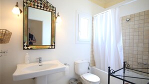 A bathroom with a white sink, toilet, and bathtub. There is a mirror with a decorative frame, a window with a white curtain, and a wicker chair. - Villa Attalus 2 (Mykonos)