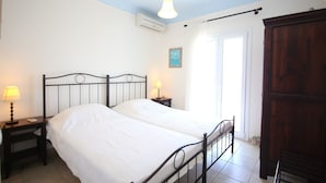 A bedroom with a bed, bedside tables, a hanging light fixture, an air conditioning unit, a wooden wardrobe, and a door leading to another room. - Villa Attalus 2 (Mykonos)