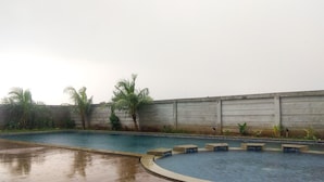 A swimming pool with a tiled deck, palm trees, and a concrete wall. - Fully Furnished Studio Apartment @ The Enviro (Cikarang)