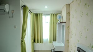 A hotel room with a bed, a bedside table, a mirror, a window with curtains, and a wall-mounted fan. - Fully Furnished Studio Apartment @ The Enviro (Cikarang)