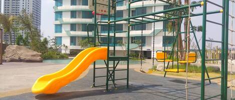 Un parque infantil con un tobogán amarillo y estructuras para escalar, rodeado por un edificio de varios pisos y ventanas.