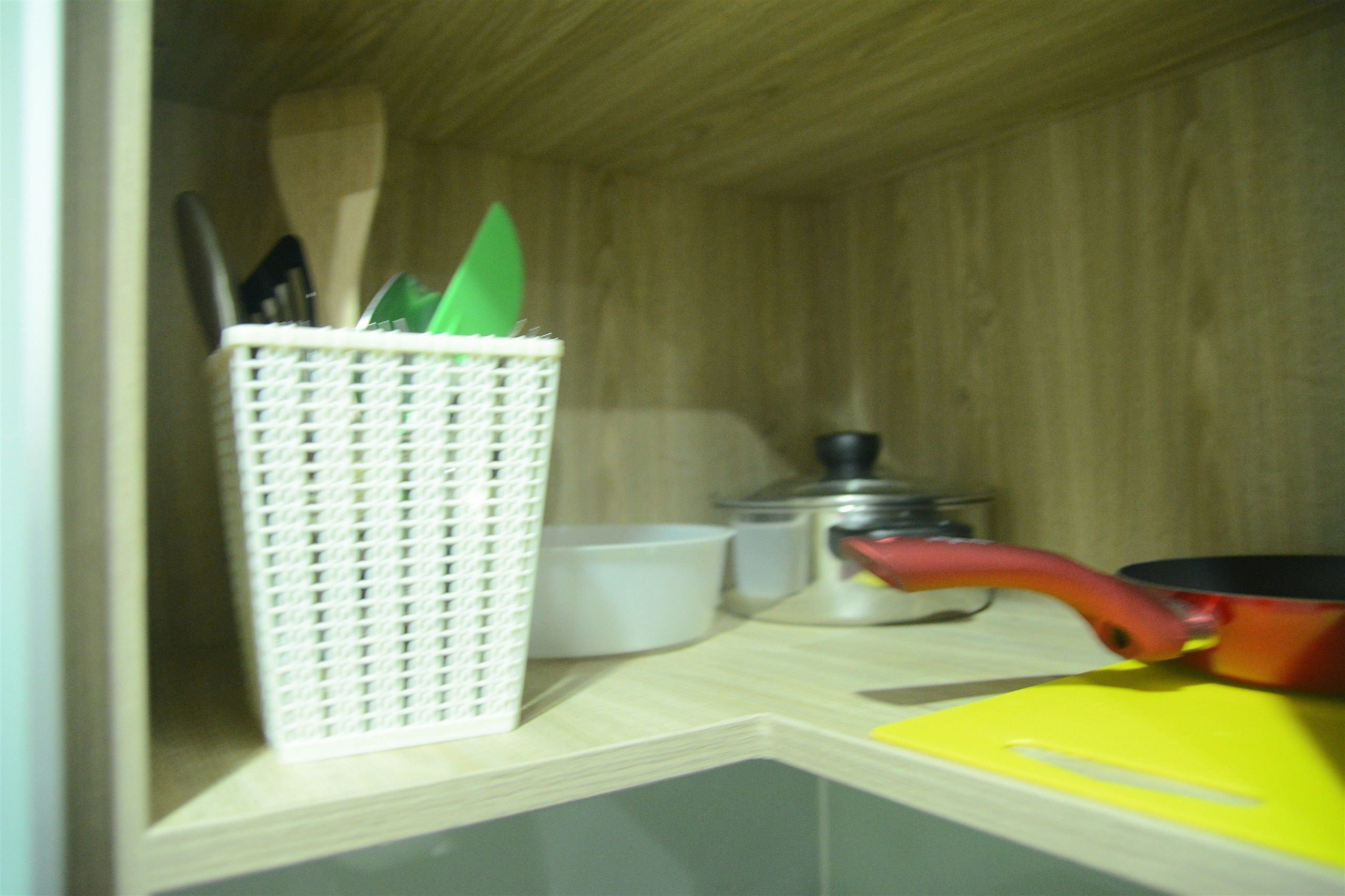 Una encimera de cocina con una cesta blanca que contiene utensilios, un bol blanco, un recipiente con tapa y una tabla de cortar amarilla.