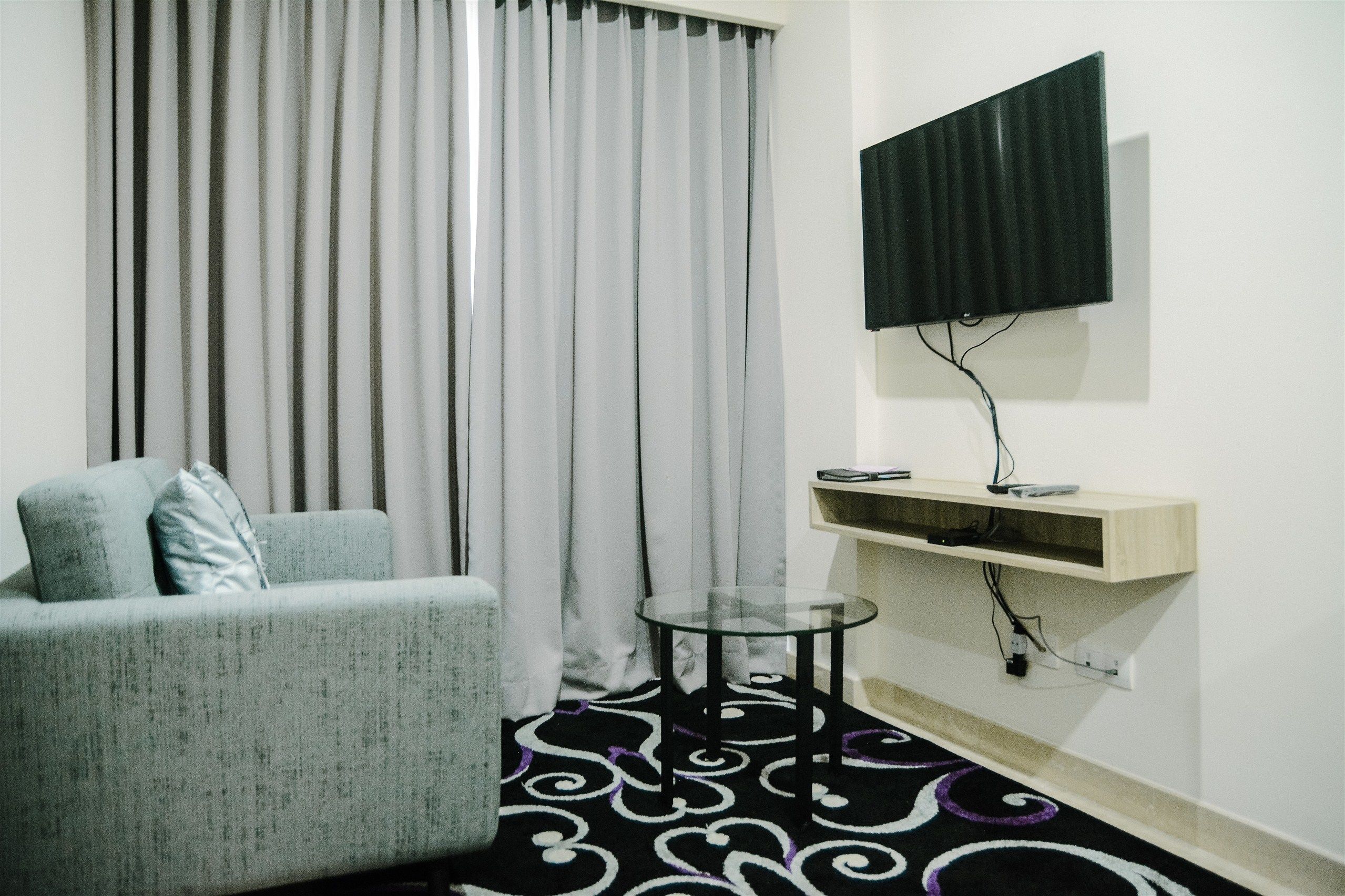 Uma sala de estar com sofá, mesa de vidro, TV na parede e prateleira com controle remoto.