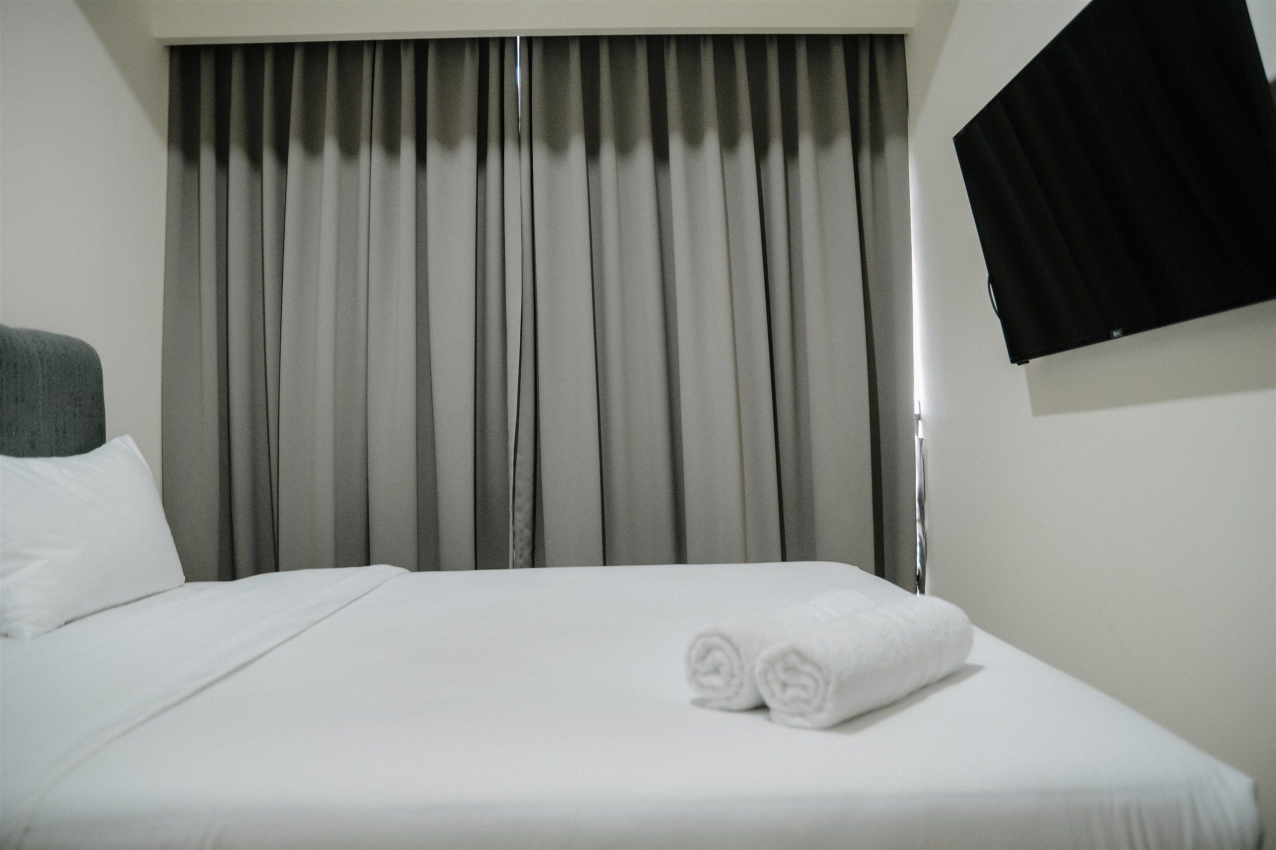 A hotel room with a neatly made bed, grey curtains, and a mounted flat-screen TV.