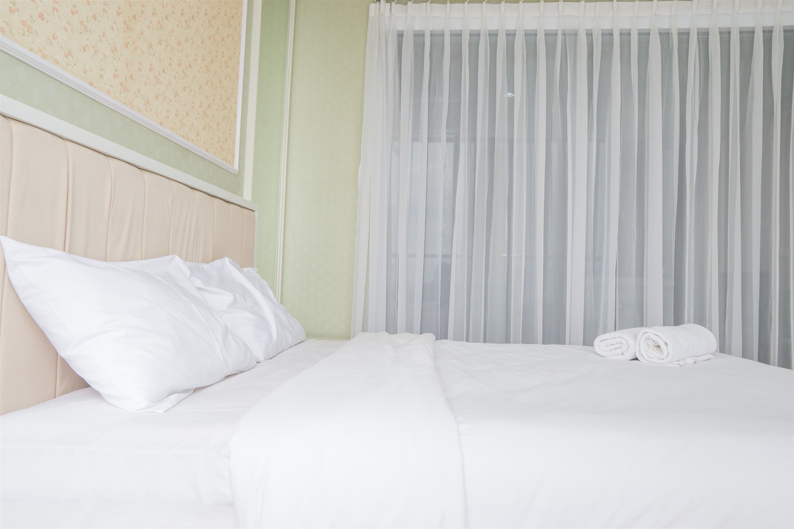 Una cama bien hecha con sábanas y almohadas blancas, una toalla enrollada a un lado y cortinas blancas traslúcidas cubriendo una ventana.