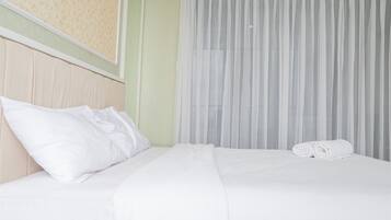 Una cama bien hecha con sábanas y almohadas blancas, una toalla enrollada a un lado y cortinas blancas traslúcidas cubriendo una ventana.