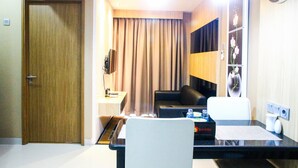 A hotel room with a wooden door, a desk with a laptop, a black leather sofa, a white chair, a clock on the wall, and a curtain. - Cozy and Minimalist 1BR @ Oasis Apartment (Cikarang)