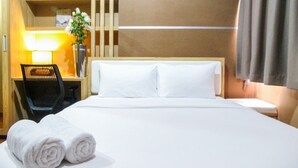 A hotel room with a neatly made bed, a desk with a chair, a lamp, and a shelf with a vase of flowers. - Cozy and Minimalist 1BR @ Oasis Apartment (Cikarang)
