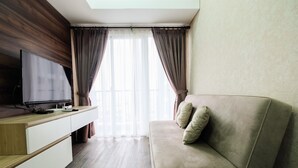 A modern living room with a flat-screen TV on a wooden console, a beige sofa with cushions, and a large window with curtains. - Graceful & Luxury 1BR Casa De Parco Apartment (Pagedangan)