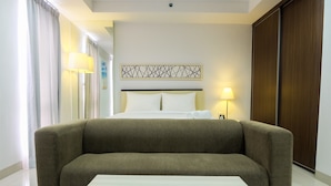 A hotel room with a bed, a sofa, a bedside table, a lamp, and a wall art piece. - Studio Room Azalea Suites Apartment (Cikarang)