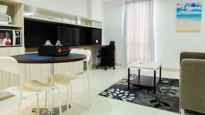 A modern living room with a round dining table, white chairs, a sofa, a TV, and a kitchen area with a microwave and coffee maker. - Studio Room Azalea Suites Apartment (Cikarang)
