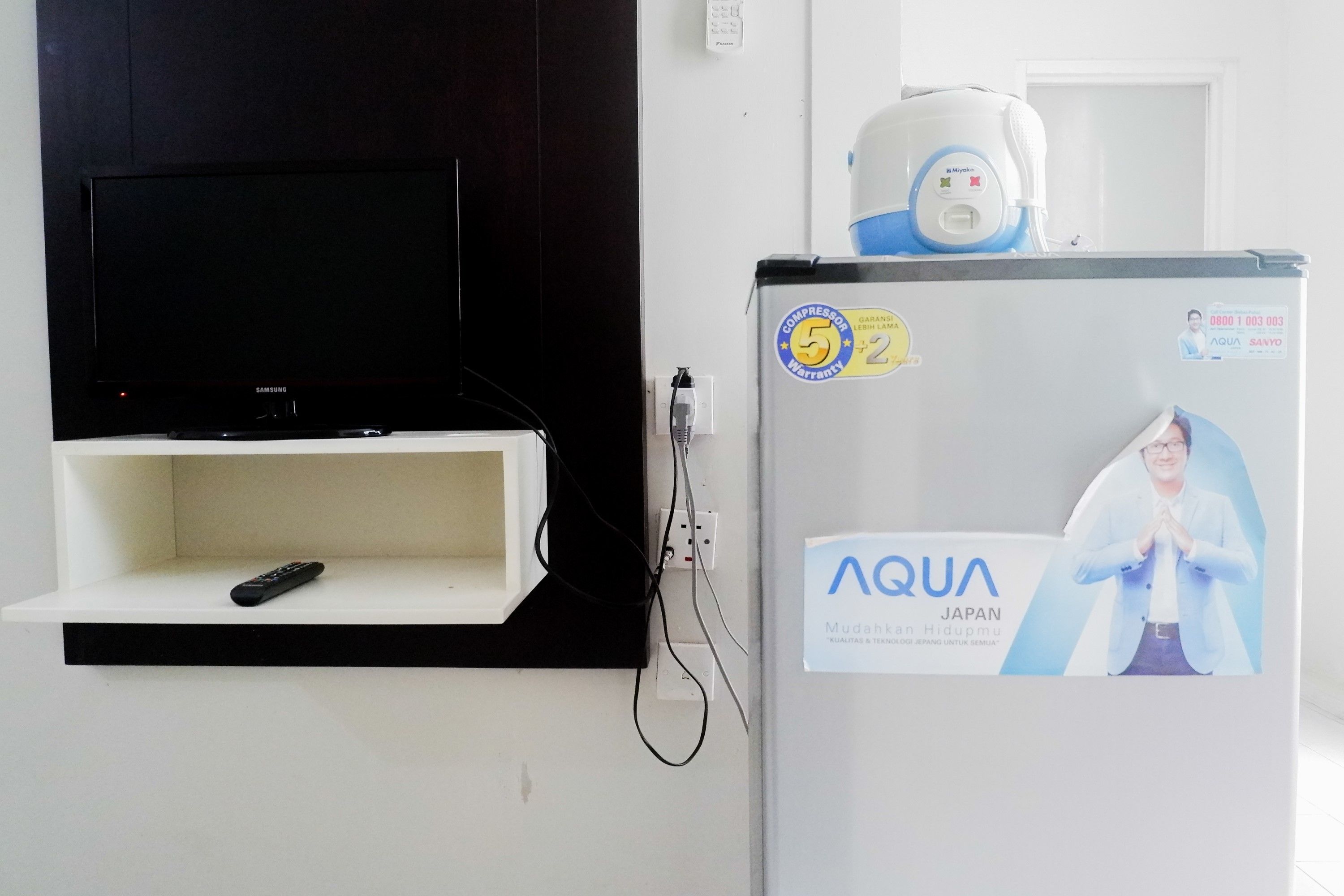 Un téléviseur Samsung fixé sur un mur noir, une étagère blanche en dessous et un réfrigérateur orné d’un autocollant AQUA et d’une image représentant une personne.
