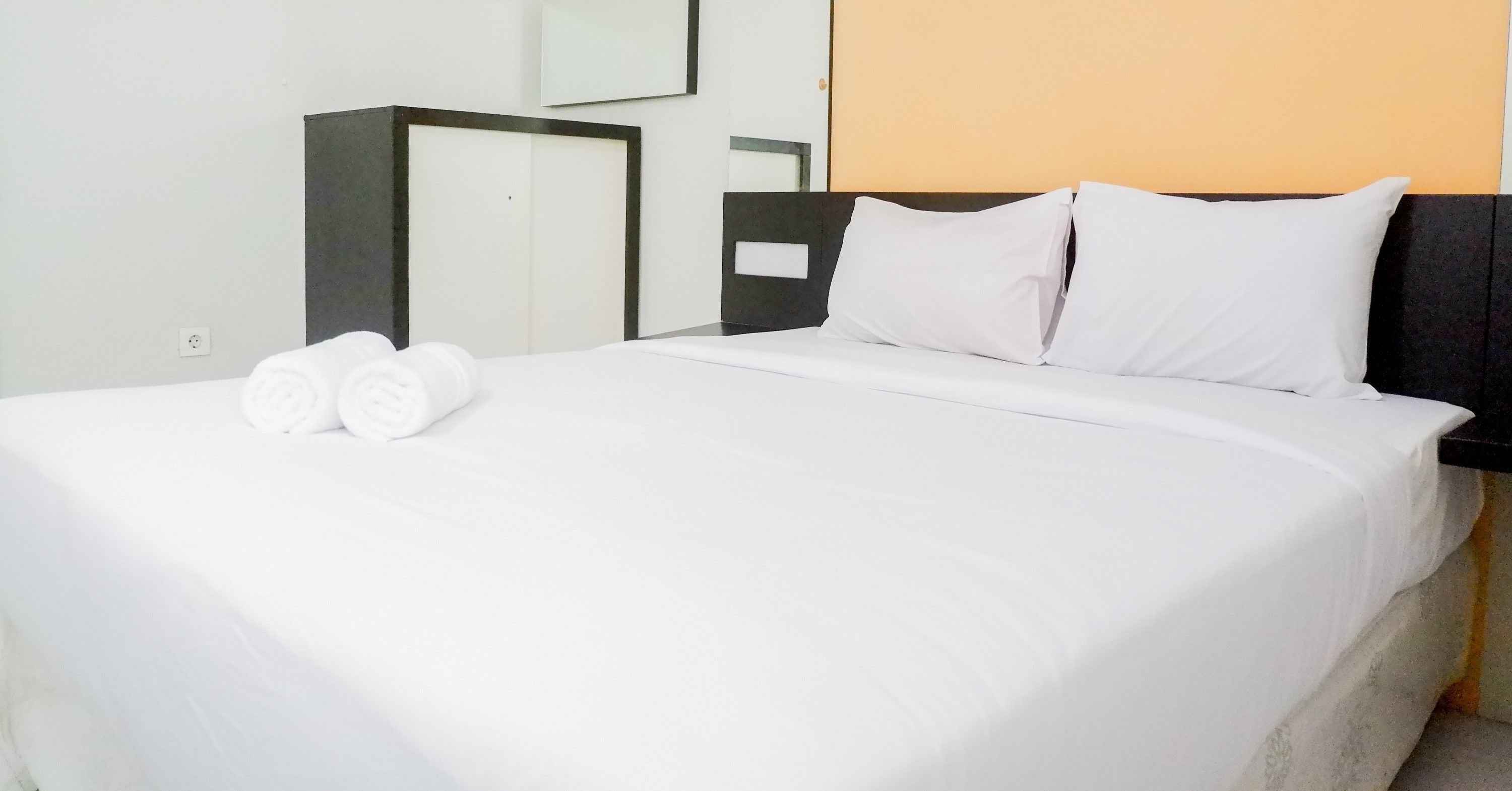 Una cama bien hecha con sábanas blancas y dos toallas enrolladas, un cabecero oscuro y un espejo fijado a la pared.