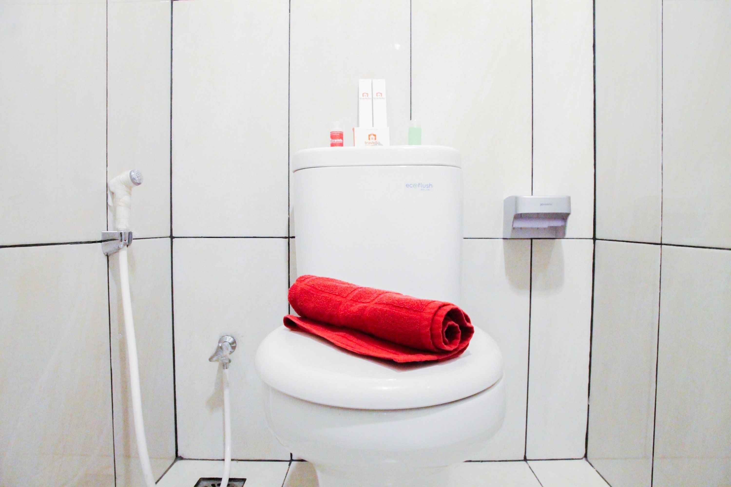 Et hvidt toilet med et rødt håndklæde ovenpå, en håndholdt brusehoved og en papirhåndklædedispenser.