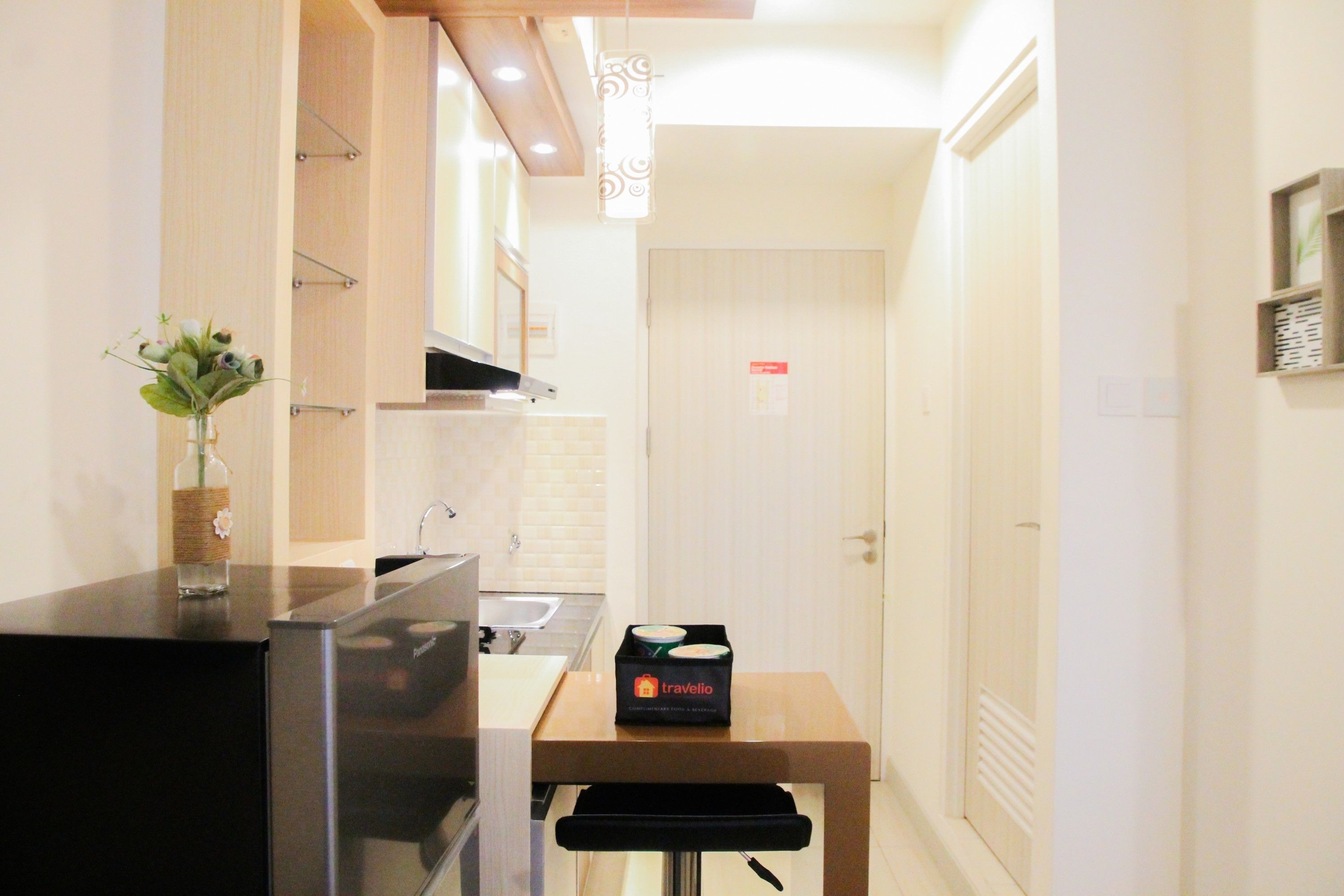 A compact kitchen with a refrigerator, a sink, and a countertop. There is a small table with a box labeled 'travelio' on it.