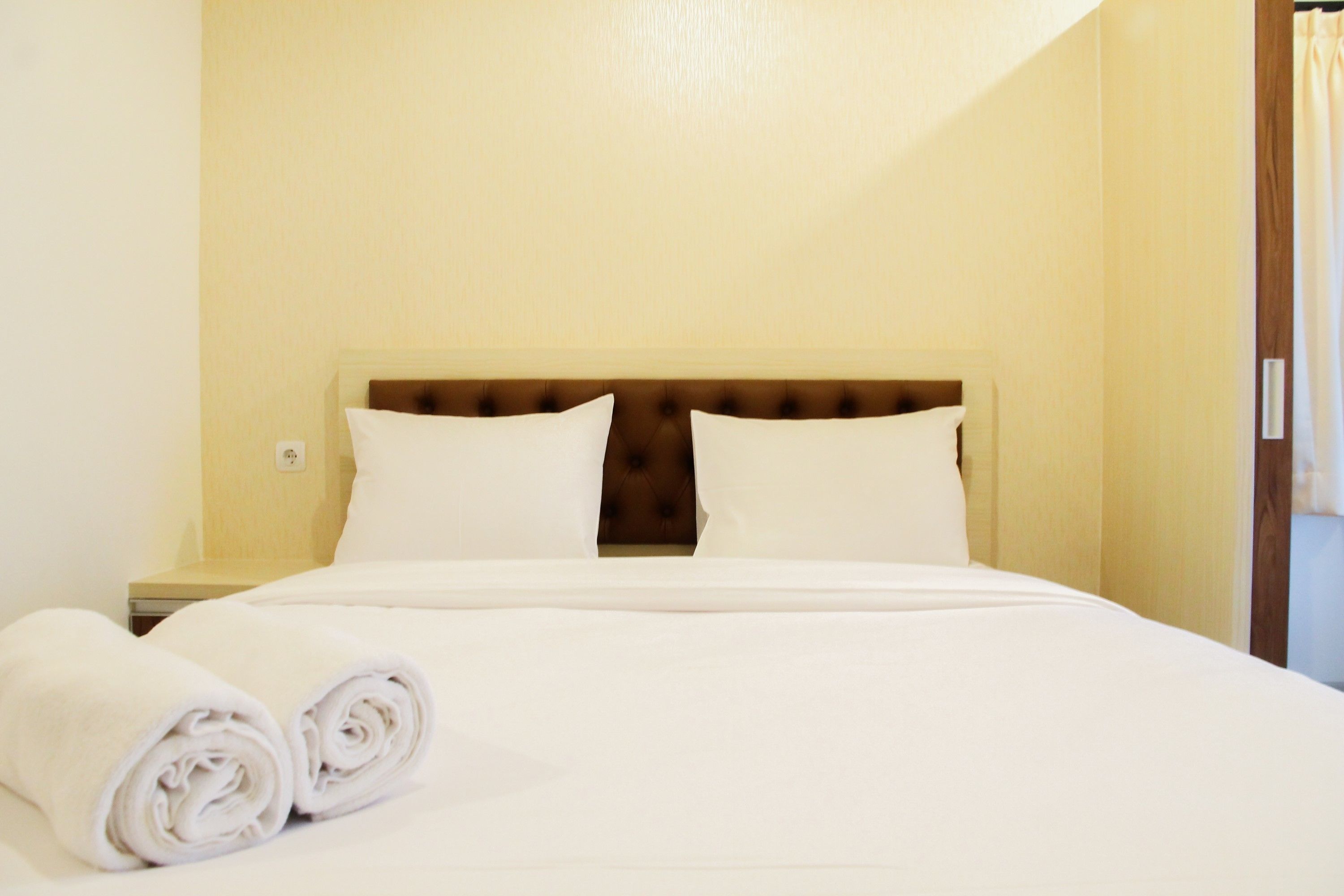 A neatly made bed with white linens and rolled towels, a wooden headboard, and a plain wall.