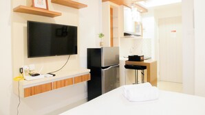 Una habitación de hotel moderna con un televisor de pantalla plana montado en la pared, un refrigerador empotrado, una pequeña cocina con estufa y lavaplatos, y una cama bien hecha con una toalla blanca.