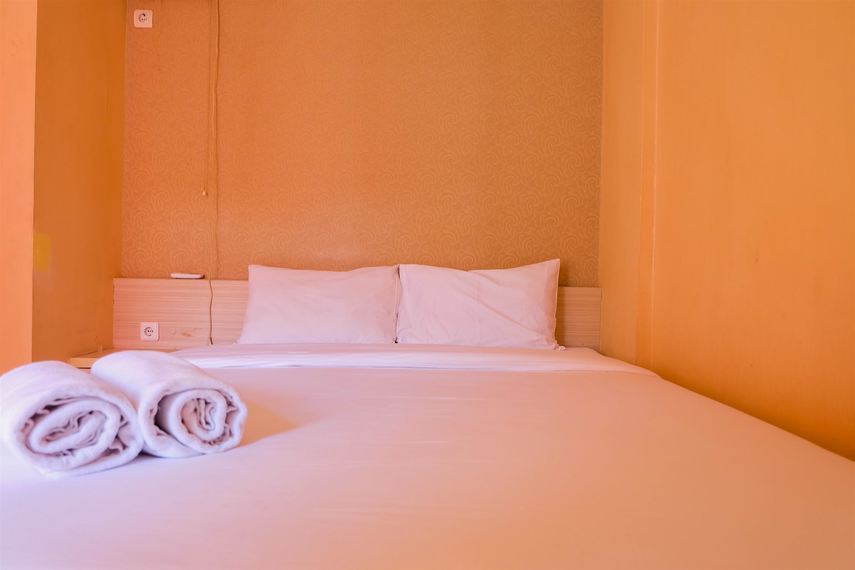 Ein ordentlich bezogenes Bett mit weißen Leinen und Kissen, ein gefaltetes Handtuch daneben und eine schlichte orangefarbene Wand.