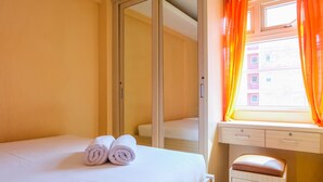 A hotel room with a bed, a small desk, a mirror, and a window with orange curtains. - Affordable Price 2BR Green Pramuka City Apartment (Jakarta)