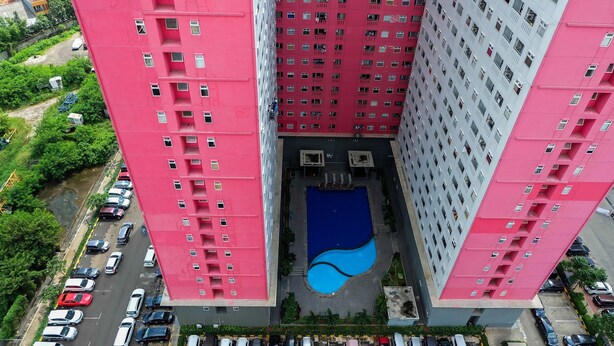 A high-rise building with multiple floors, a central courtyard with a swimming pool, and a parking area with several cars. - Affordable Price 2BR Green Pramuka City Apartment (Jakarta)