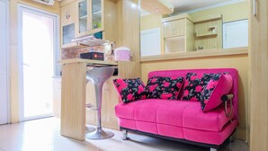 A bright room with a pink sofa, a small kitchen area, and a door leading to an outdoor area. - Affordable Price 2BR Green Pramuka City Apartment (Jakarta)