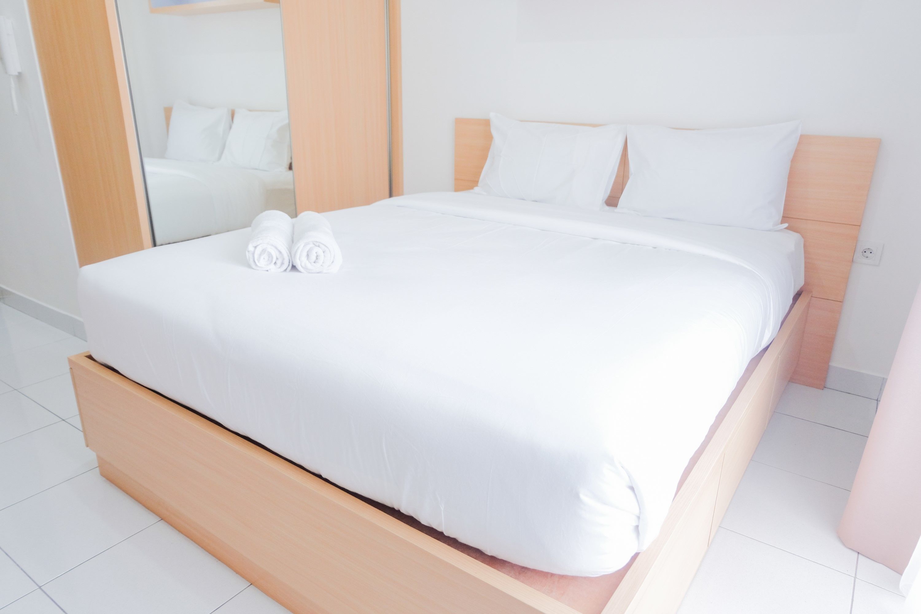 A neatly made bed with white linens and two rolled towels, a wooden headboard, and a large mirror on the wall.