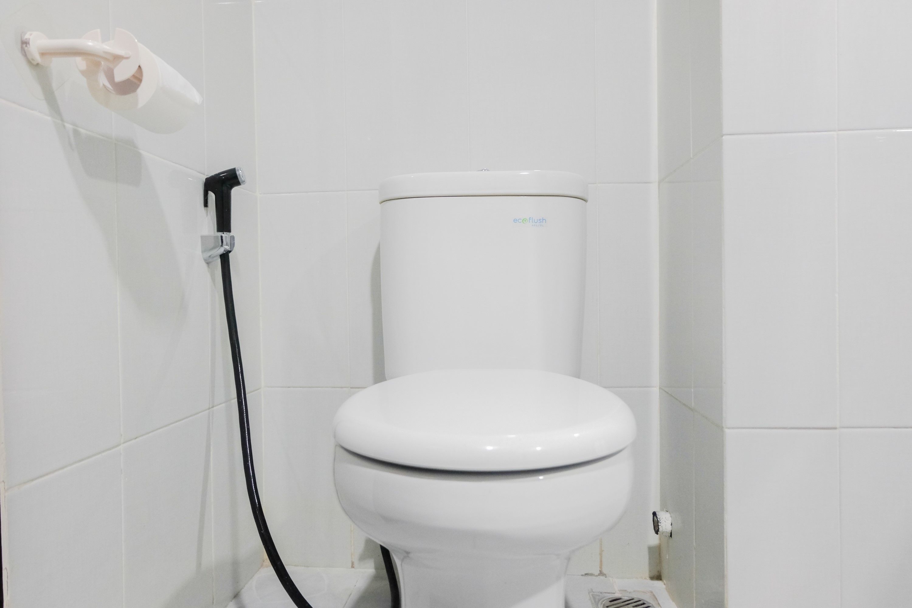 A white toilet with a black hose attached to the wall, a white toilet paper holder, and a white tiled wall.